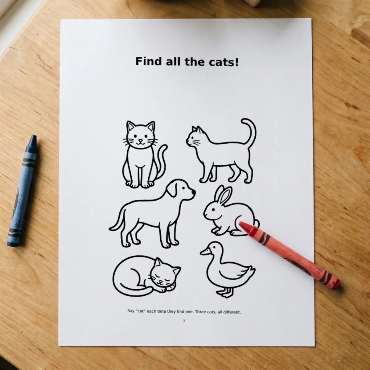 The My First Busy Book page “Find all the cats!” printed and lying on a wooden table, a red crayon across its corner.