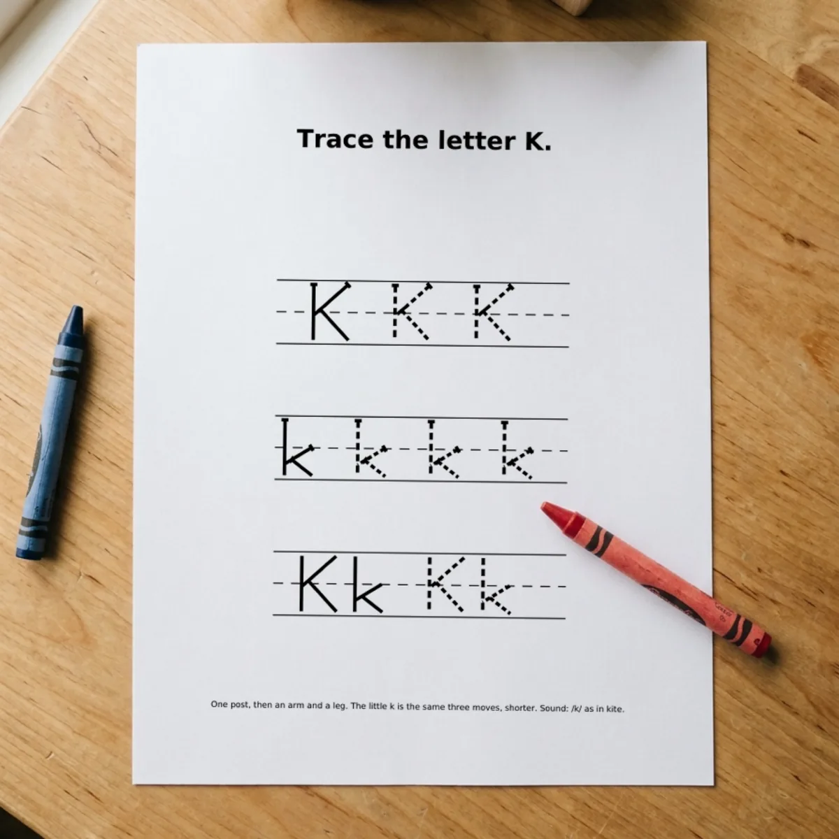 The Letters & Sounds page “Trace the letter K.” printed and lying on a wooden table, a red crayon across its corner.