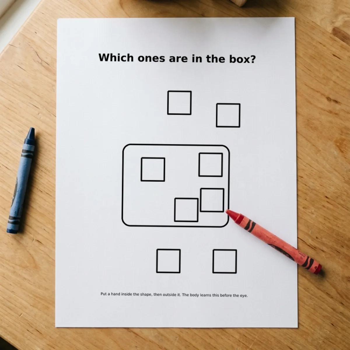 The Shapes & Sizes page “Which ones are in the box?” printed and lying on a wooden table, a red crayon across its corner.