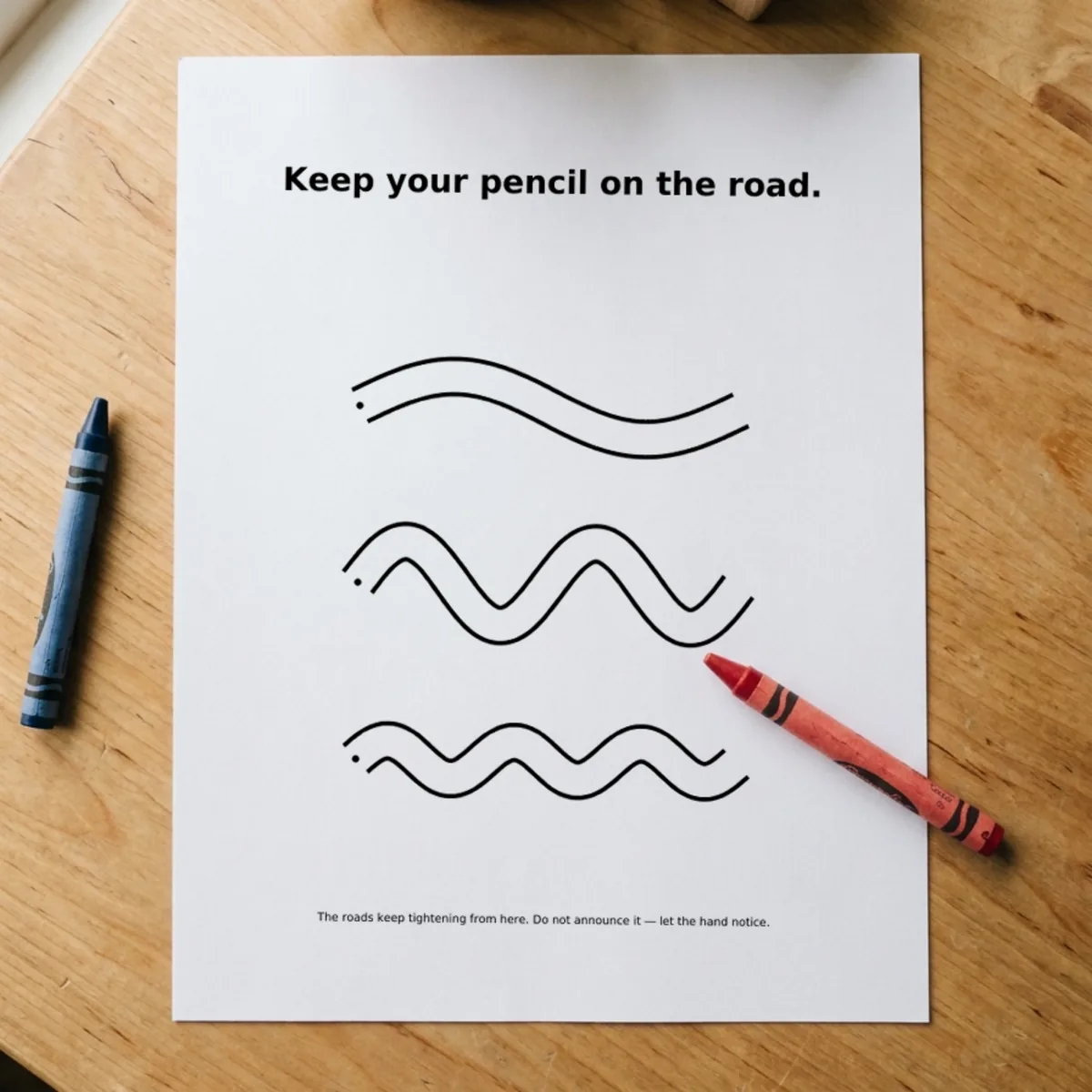 The Prewriting page “Keep your pencil on the road.” printed and lying on a wooden table, a red crayon across its corner.
