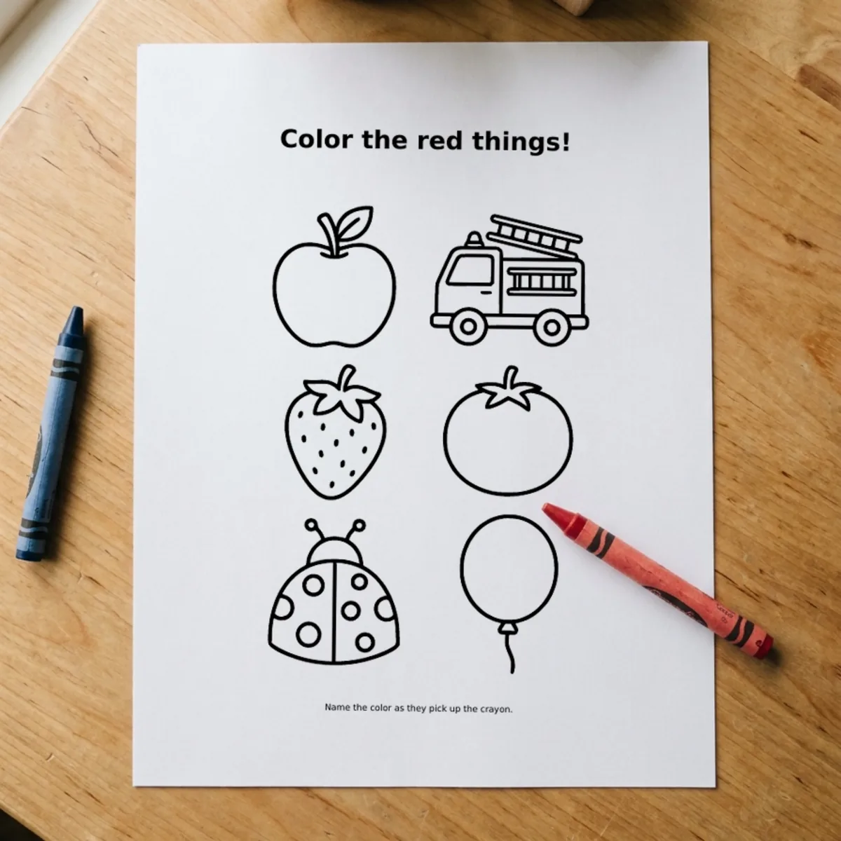 The Toddler Activity Bundle page “Color the red things!” printed and lying on a wooden table, a red crayon across its corner.