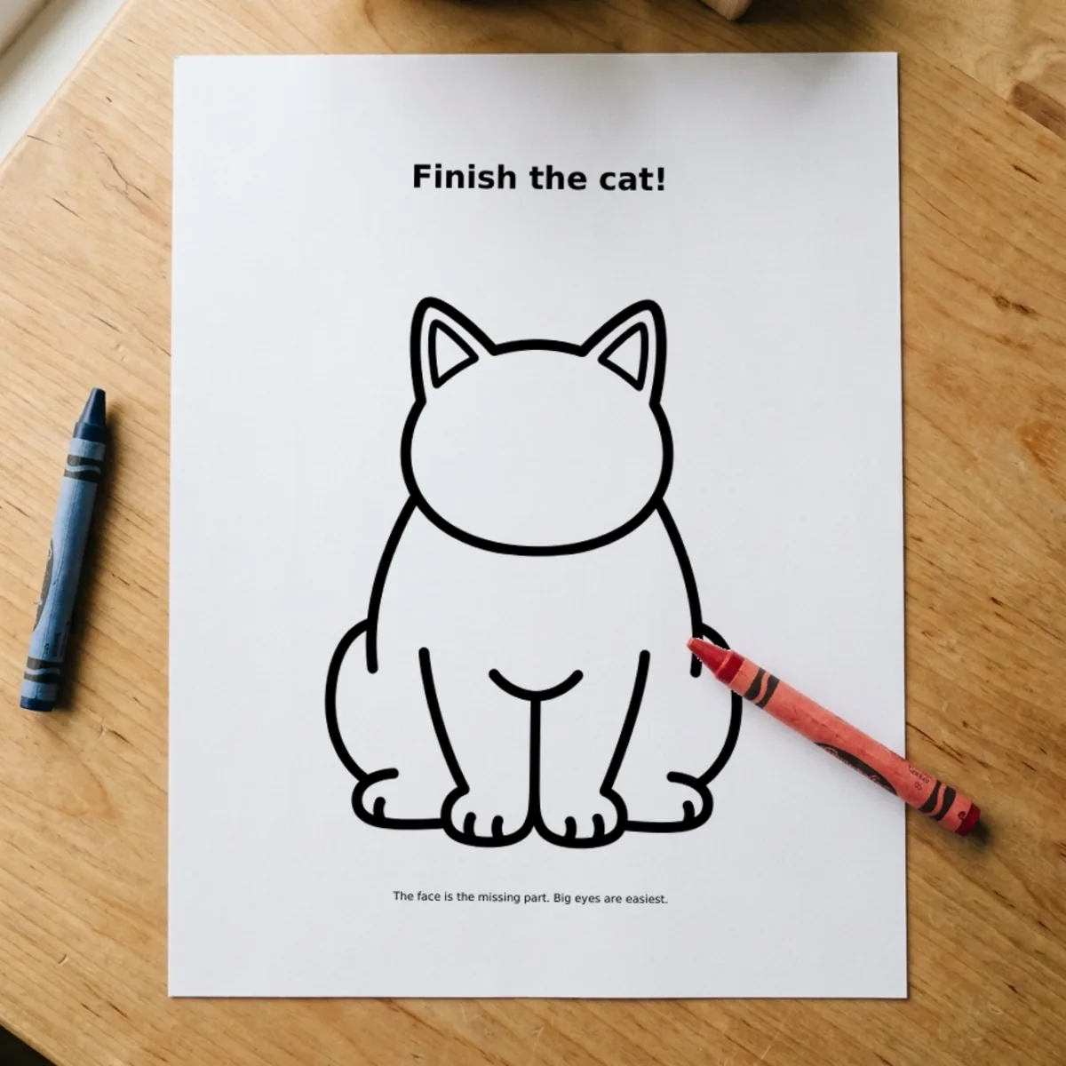 The Drawing Prompts page “Finish the cat!” printed and lying on a wooden table, a red crayon across its corner.