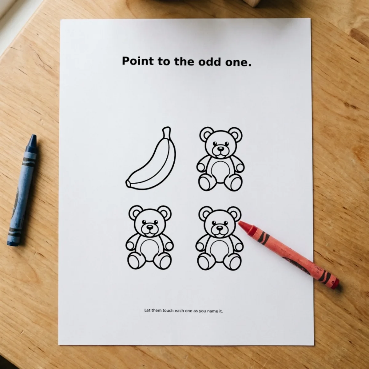 The Matching Games page “Point to the odd one.” printed and lying on a wooden table, a red crayon across its corner.