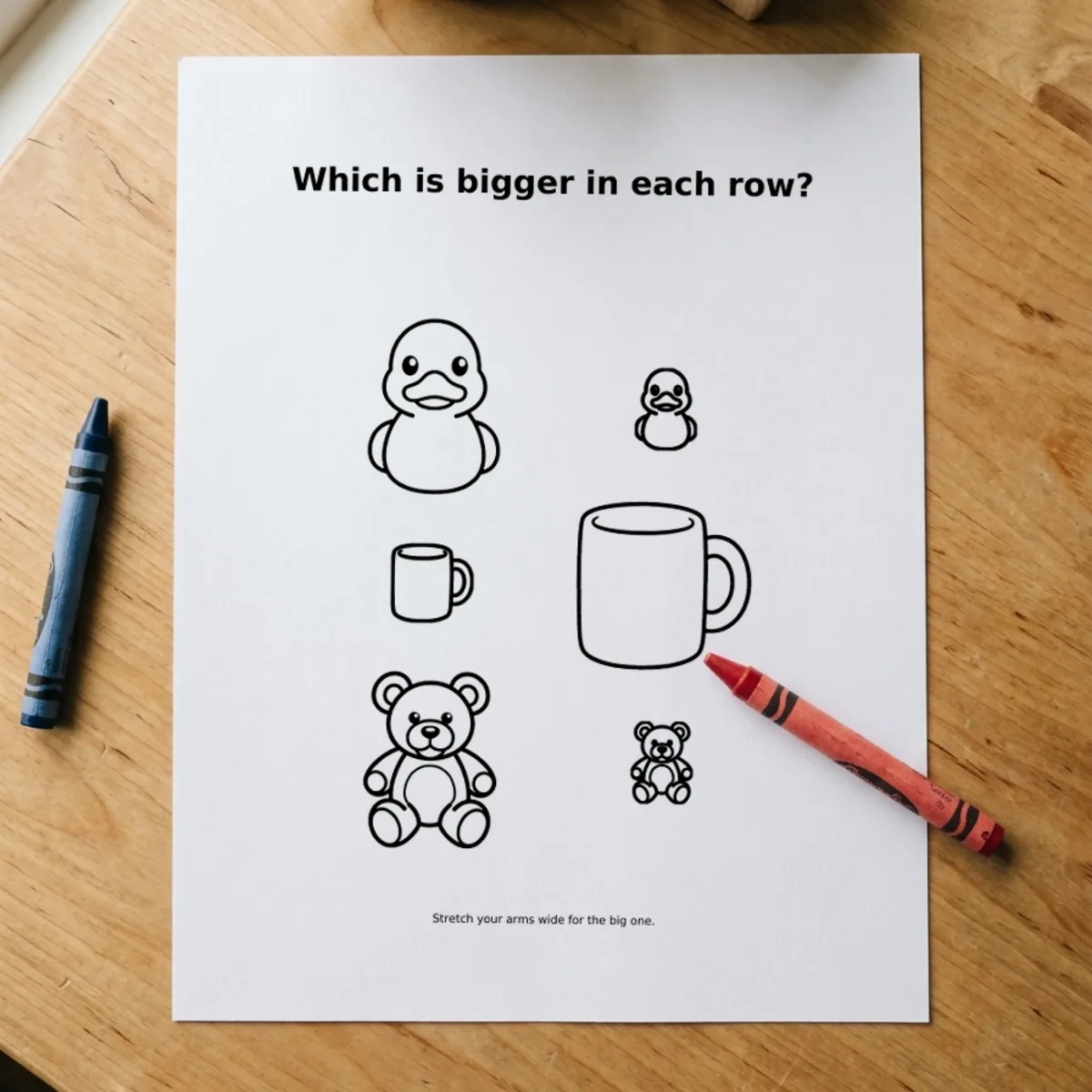 The Shapes & Counting page “Which is bigger in each row?” printed and lying on a wooden table, a red crayon across its corner.