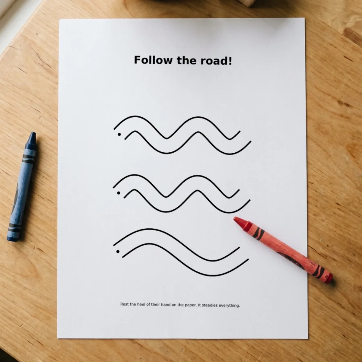 The Tracing Practice page “Follow the road!” printed and lying on a wooden table, a red crayon across its corner.